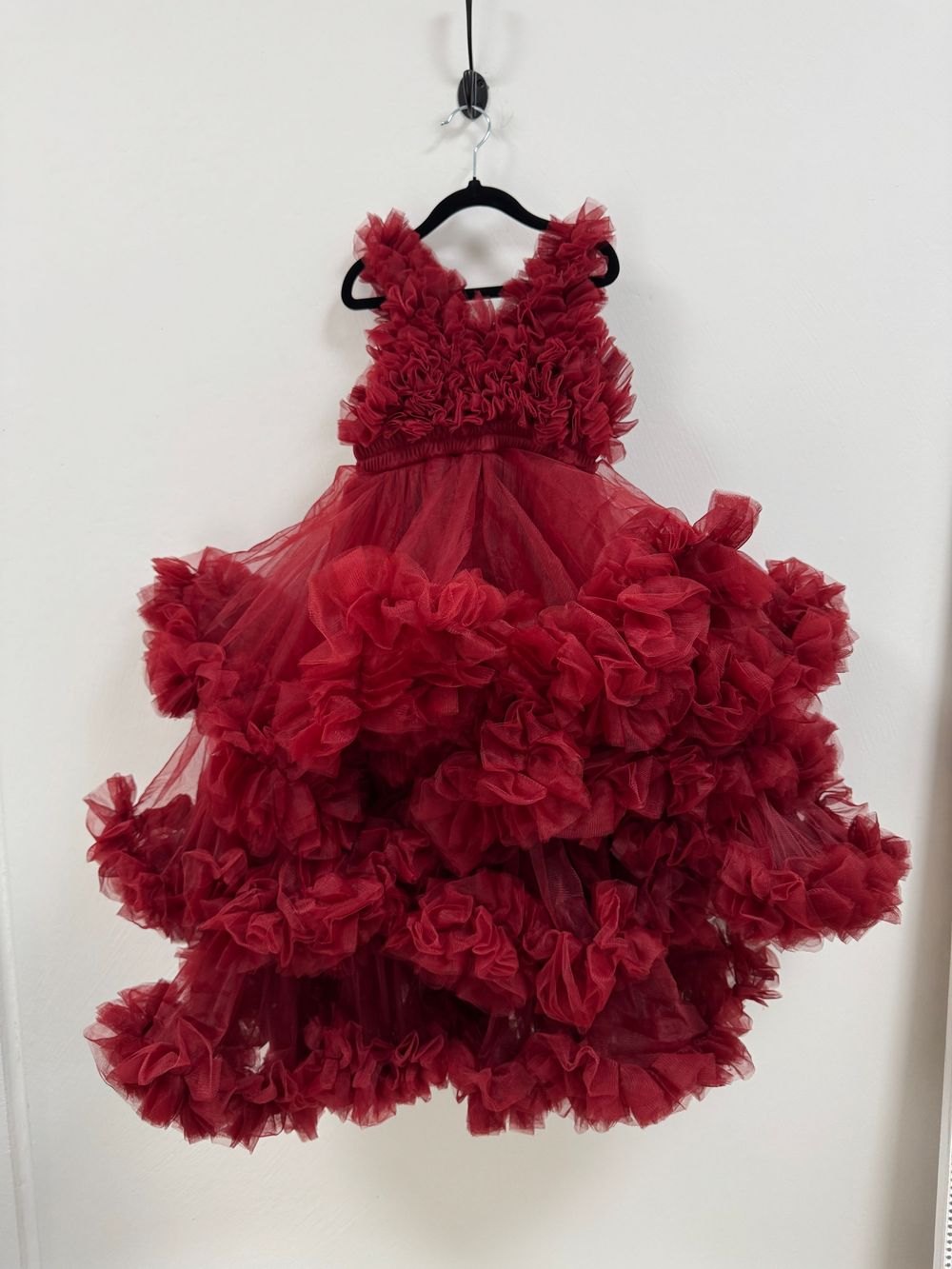 Luxurious red tulle dress with voluminous ruffled skirt and floral bodice details hangs elegantly against white wall.