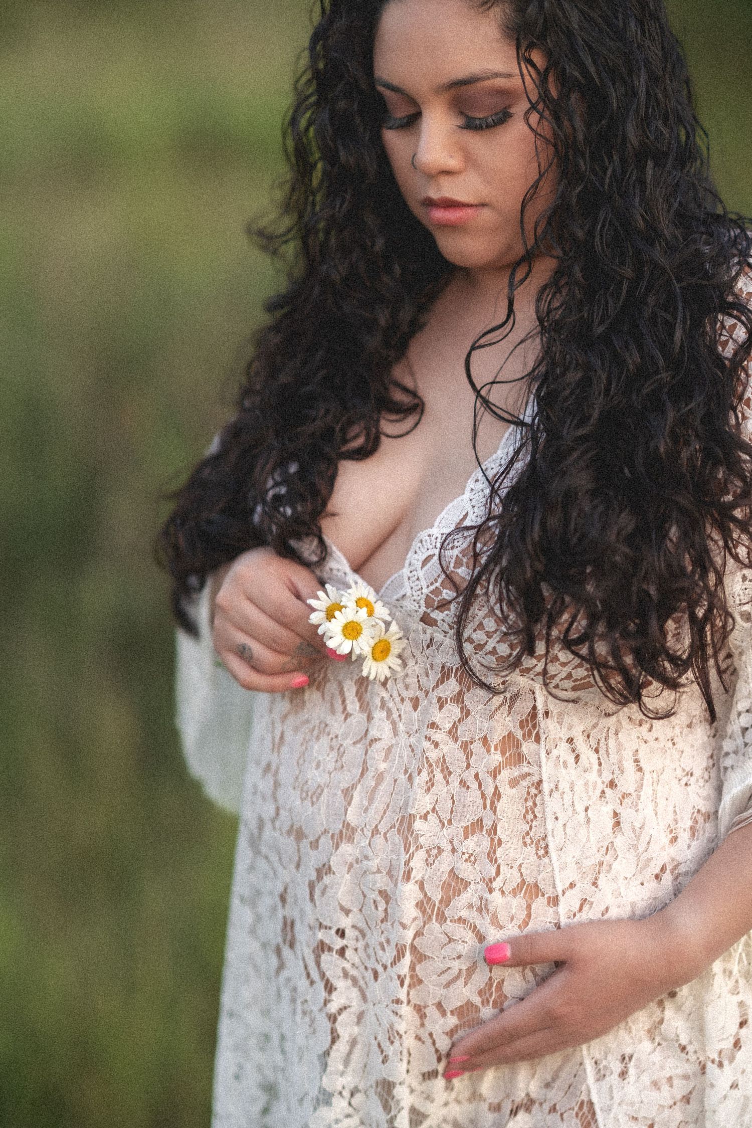 Long wavy dark hair and white lace dress holding small daisy flowers in a dreamy outdoor portrait scene.