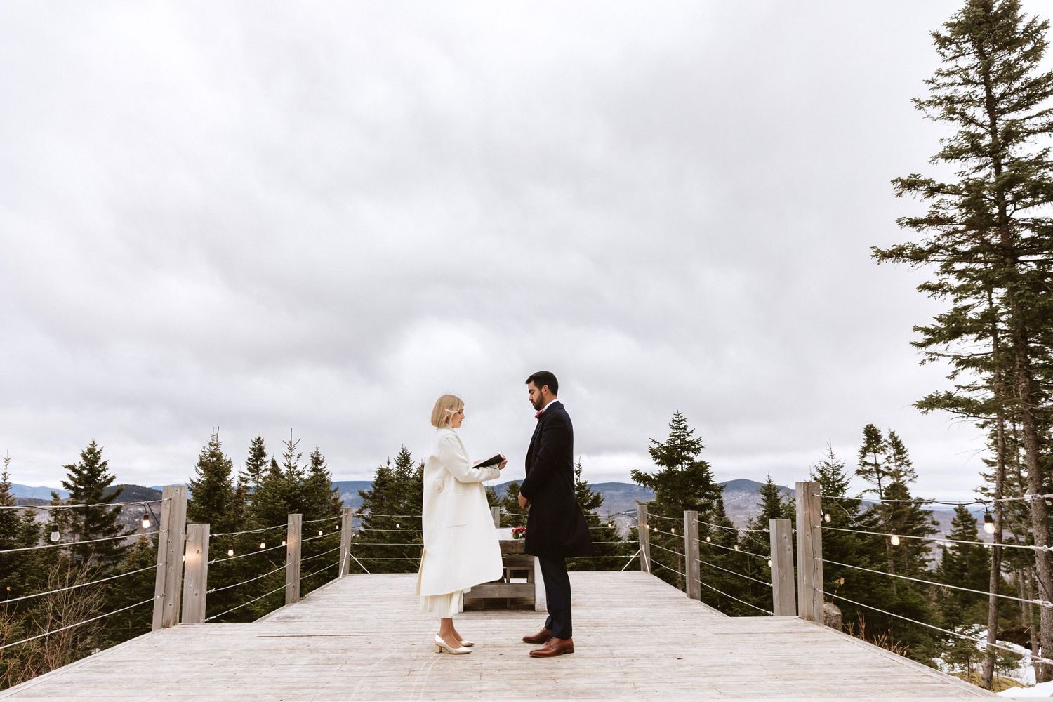 A couple shares a romantic moment on a wooden deck overlooking evergreen trees on their wedding day.