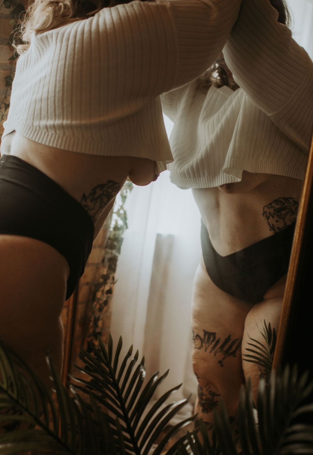 Artistic boudoir photograph showing silhouettes in soft light with palm fronds and cropped white sweaters.