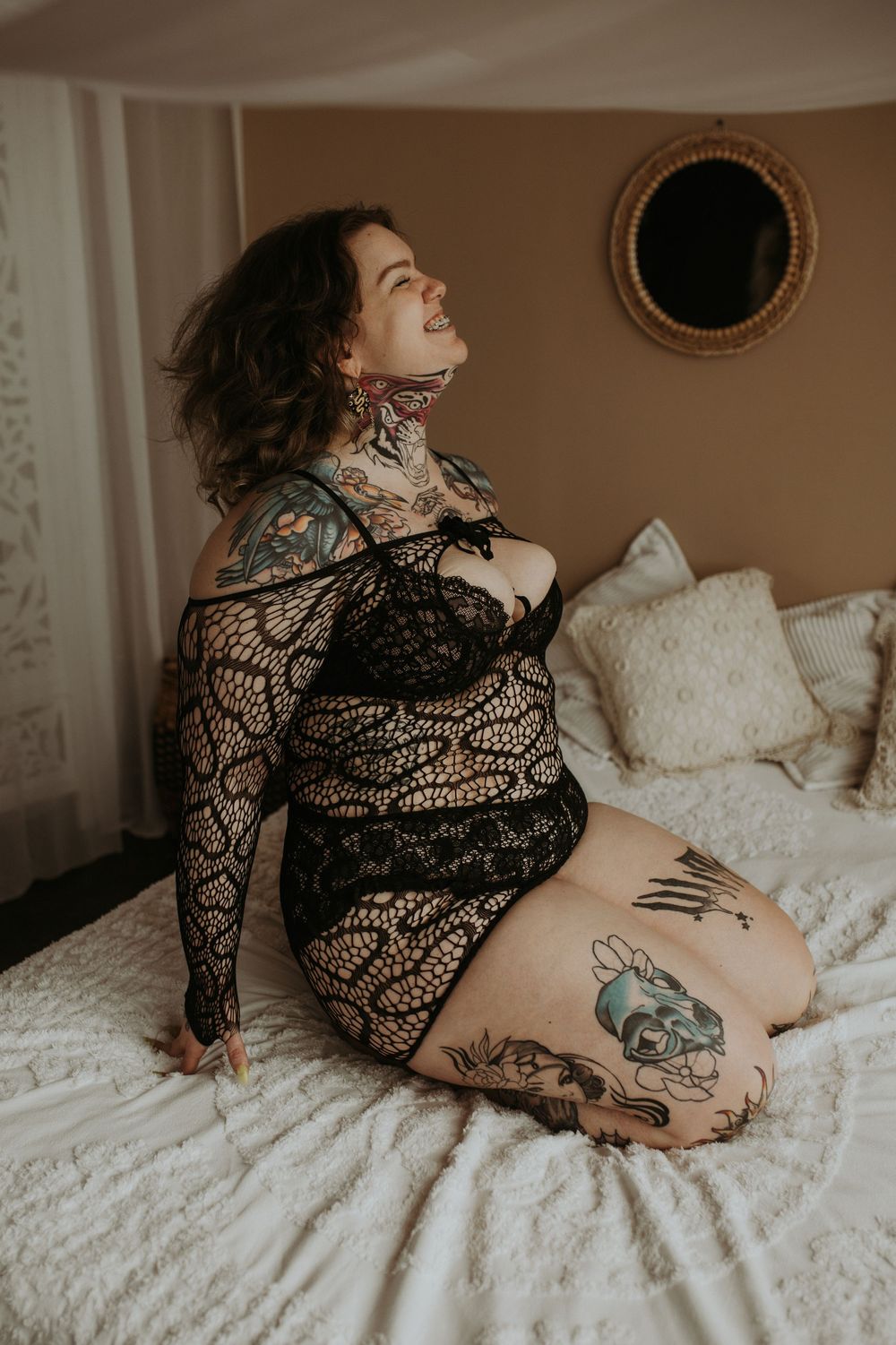 A person in black lace lingerie poses on white bedding, showing artistic tattoos in a moody boudoir setting.