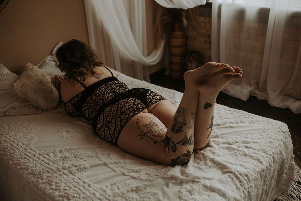 A person in black lace lingerie lying on a white bed with sheer curtains in moody lighting.