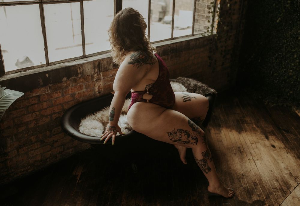 A woman in lingerie sits near a window on a wooden floor in moody, artistic lighting.