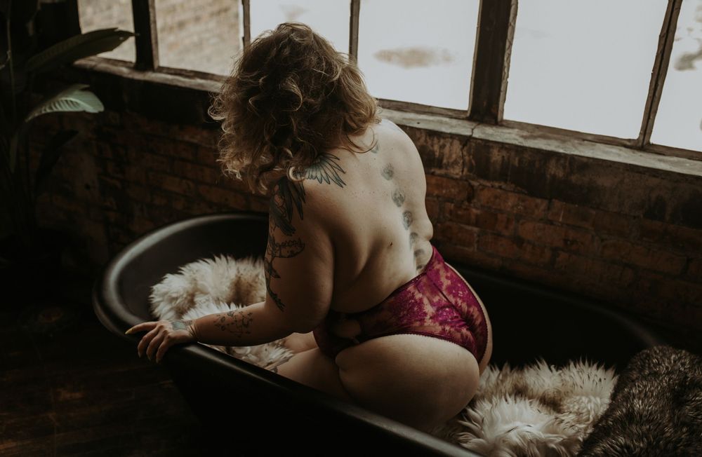 Person seated on dark floor near exposed brick wall and window with sheepskin rug.