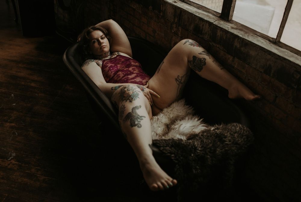Moody boudoir photograph of someone reclining on vintage couch next to window in dark room with soft natural lighting.