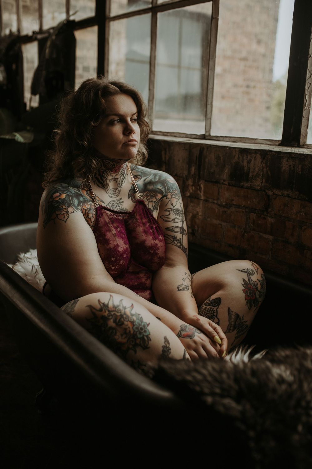 Moody indoor portrait showing a person in burgundy dress with tattoos sitting by window in dark atmospheric lighting.