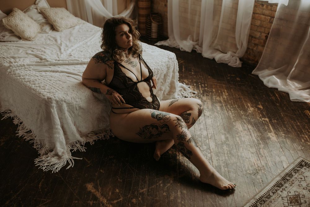 Figure in black lingerie poses against white sheets while natural light pours in through window.