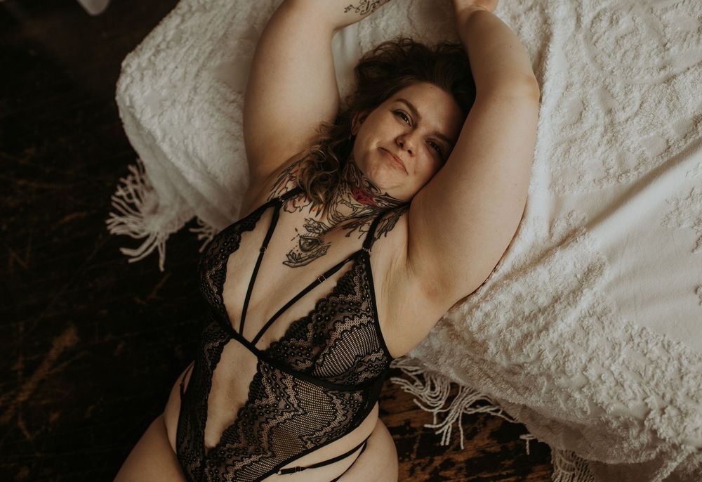 Figure in lace bodysuit reclining on white textured bedding with dramatic lighting.