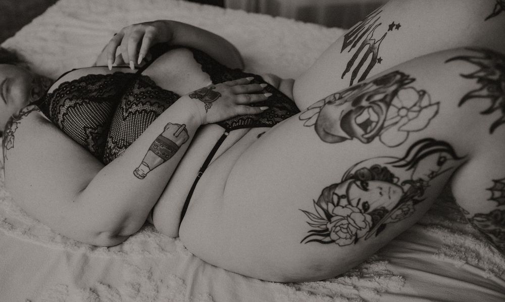 Black and white photo showing intricate tattoo artwork on torso including roses and decorative designs while laying on bedding.