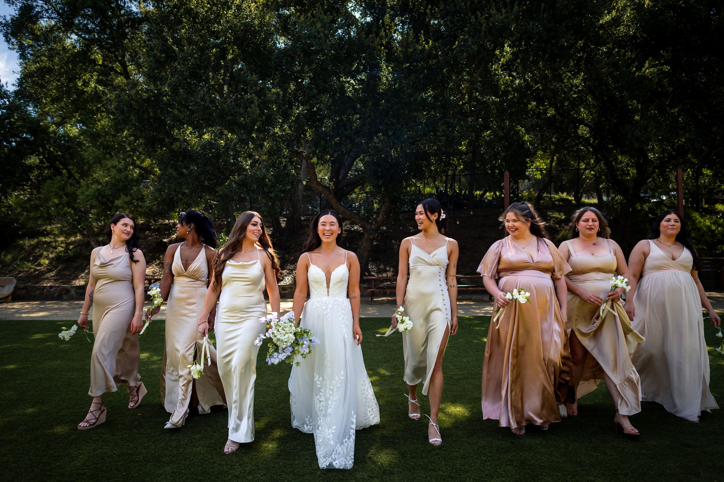 Wedding party in white and neutral dresses posing outdoors in garden setting.
