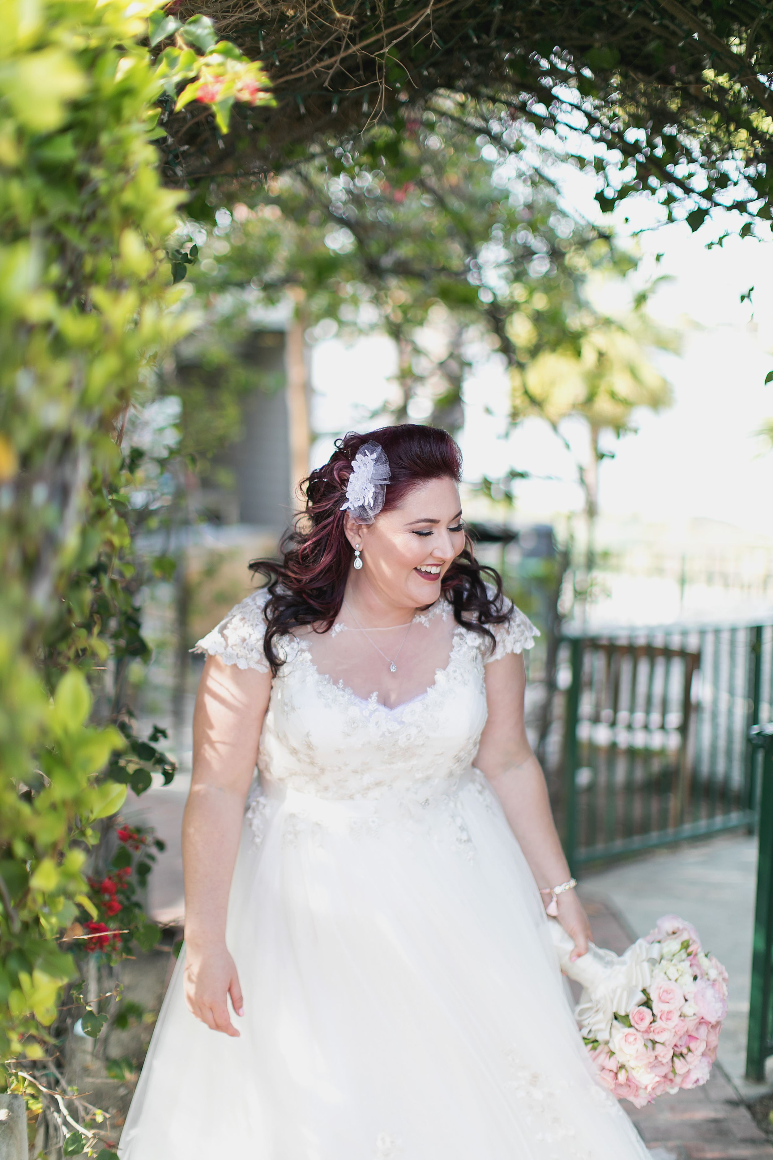 A bride in a white lace dress with curled dark hair holds a pink bouquet in a garden setting.