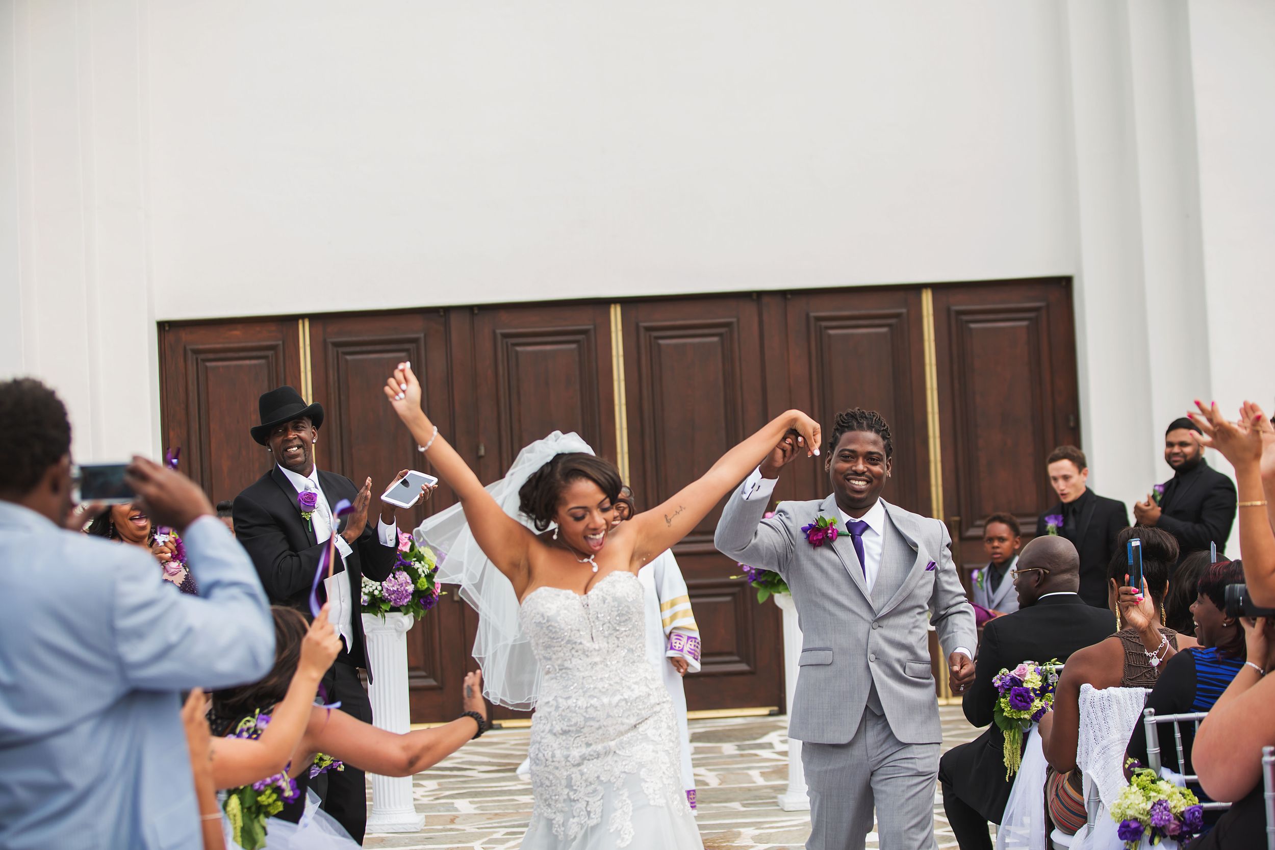 Newlyweds celebrate with raised arms while exiting church as guests cheer.