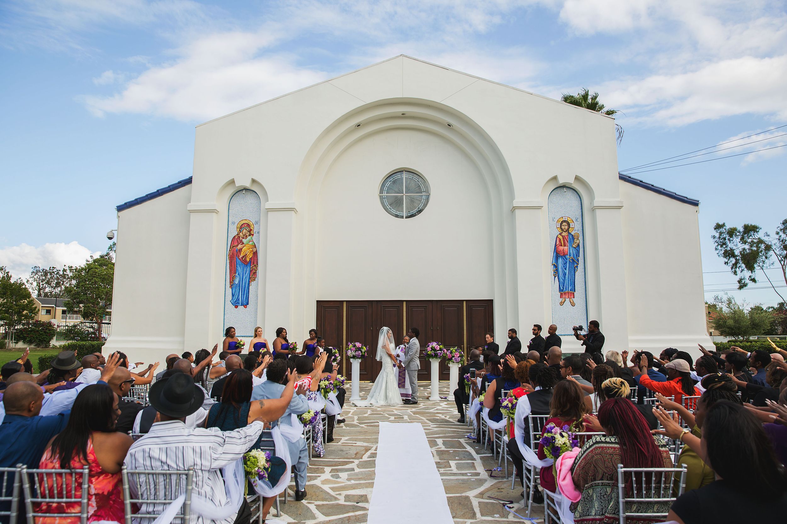 Large crowd watches wedding ceremony outside white church with stained glass windows and arched entrance.