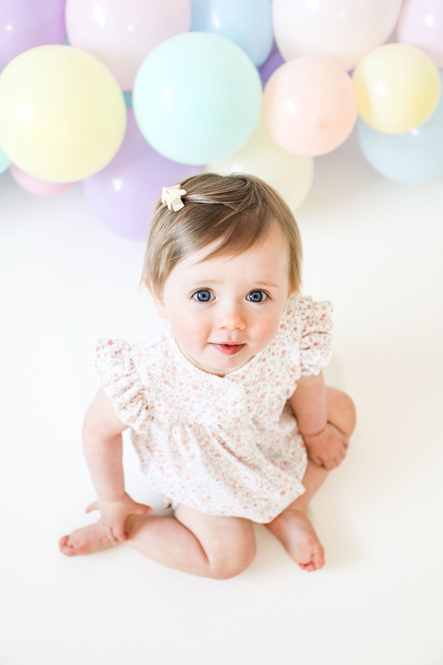 A baby in a floral lace romper sits on a white backdrop with colourful pastel balloons arranged above, during birthday shoot in Nottingham