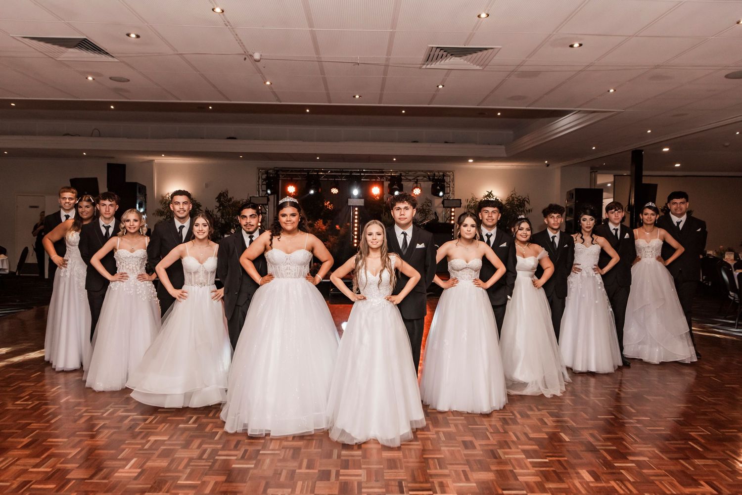 Beautiful wedding party in formal attire stands together on a wooden dance floor in an elegant ballroom.
