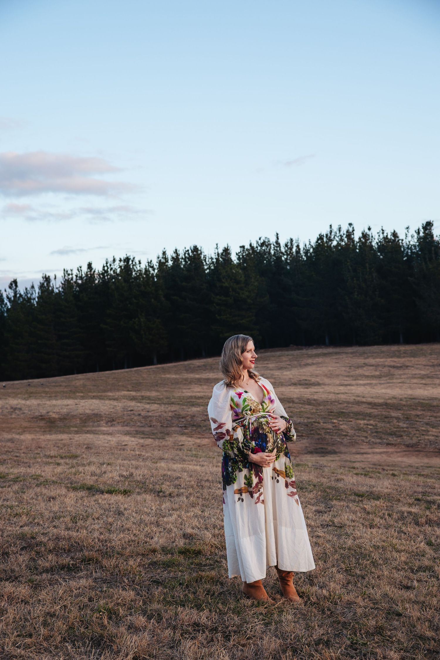 A person in a floral dress stands in a brown field at dusk with pine trees silhouetted against the sky.