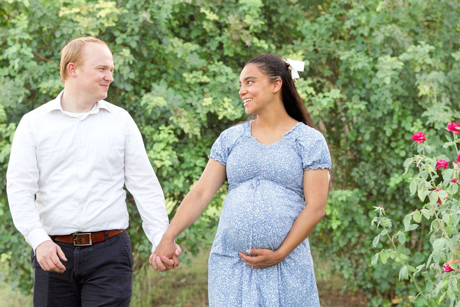 Couple holding hands smiling at each other while woman holds her baby bump in the middle of a garden in Mesa.