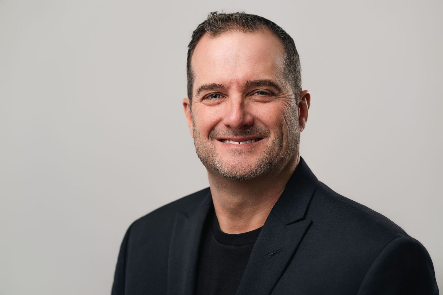 Professional headshot of a smiling middle-aged man in a black blazer against a light gray background.