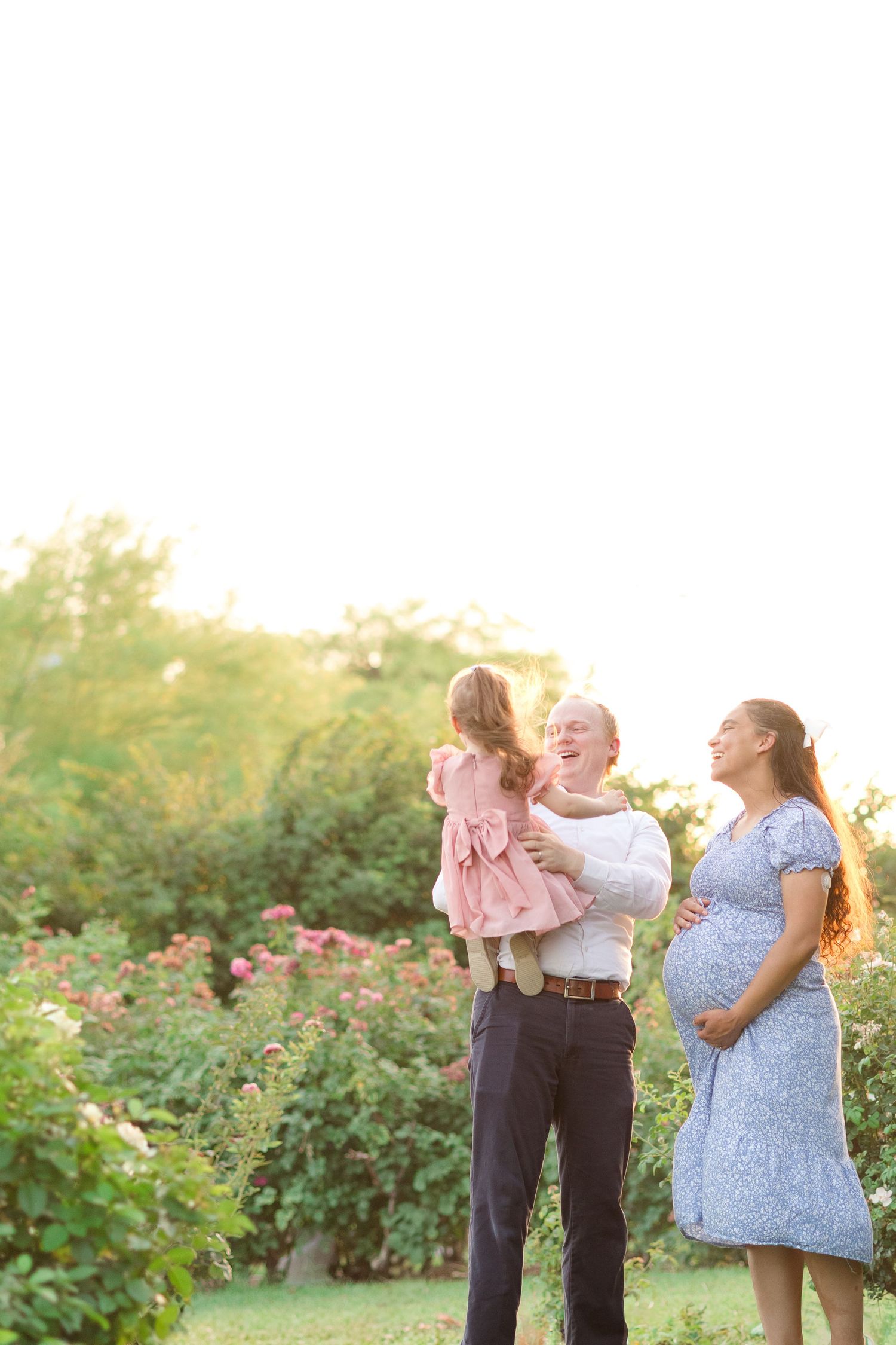 Family of 3, maternity photos,in a rose garden with dad holding and smiling at toddler daughter and mom cradling pregnant belly next to them
