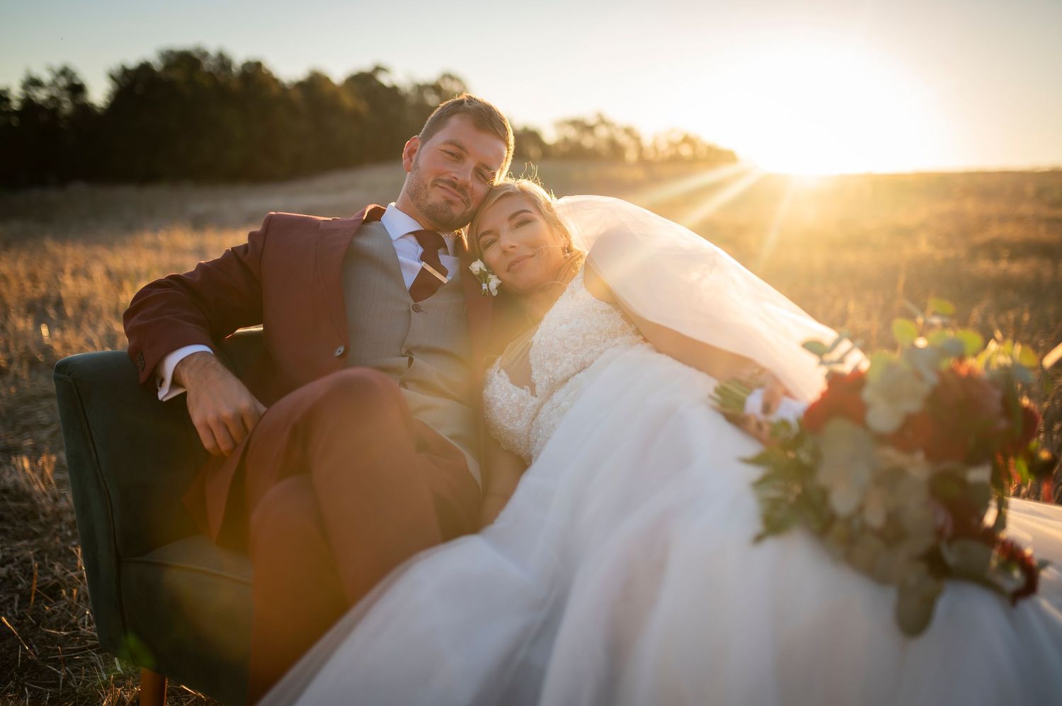 Wedding couple embraces at sunset in a romantic outdoor setting with golden sunlight filtering through the trees.
