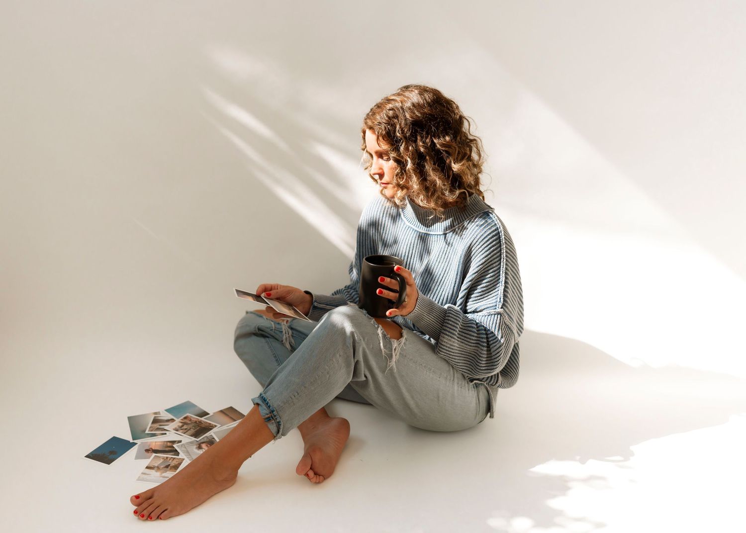 Someone sits barefoot on a white floor looking at photos while holding a coffee cup in natural window light.