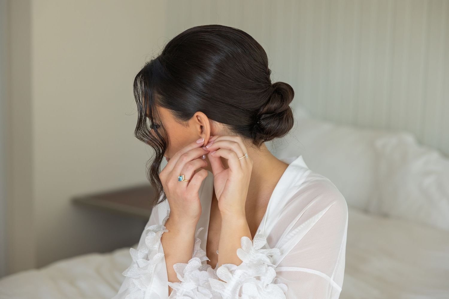 A person in a white sheer robe puts on earrings while preparing for a special occasion, with an elegant updo hairstyle.