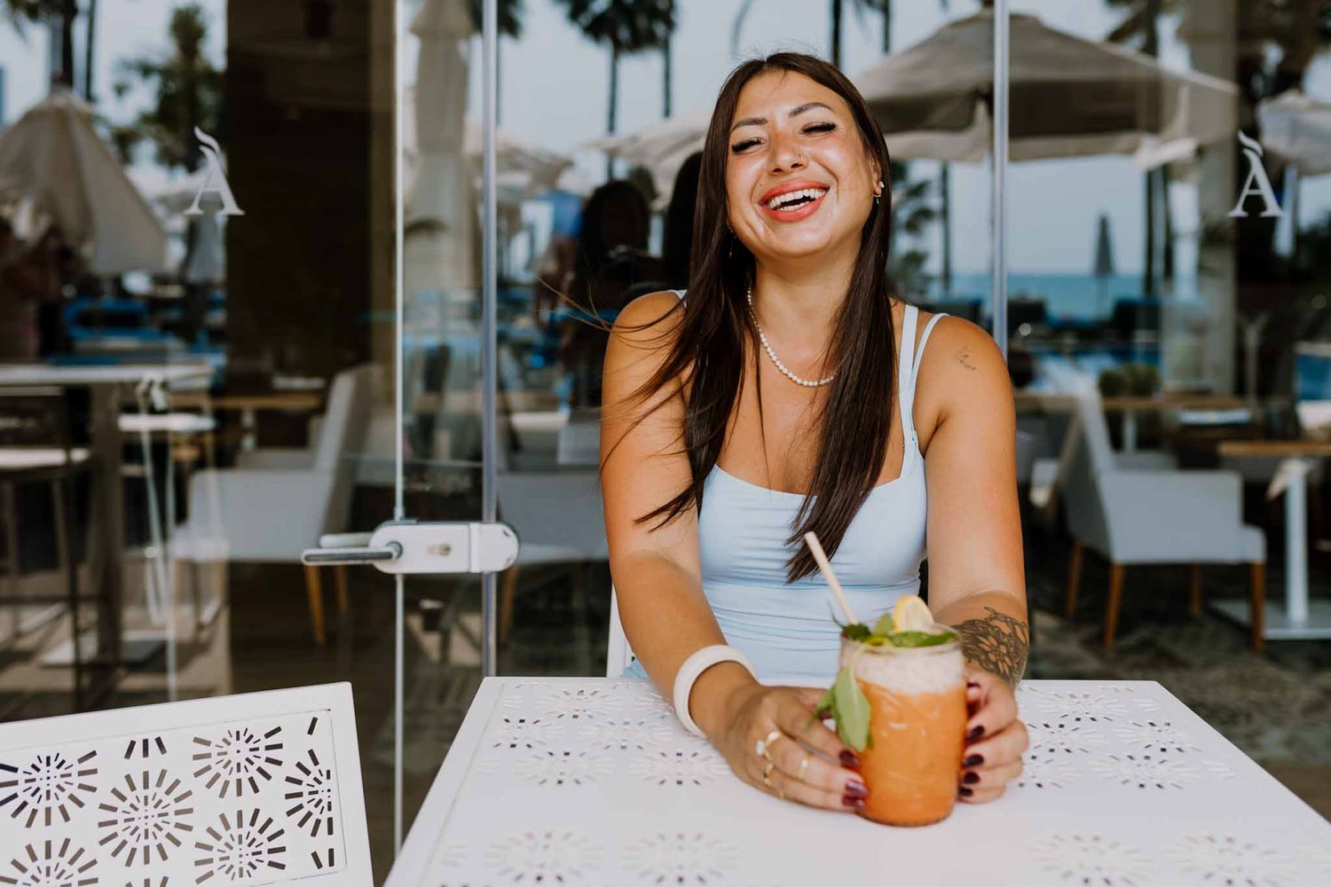 Smiling woman in a blue dress enjoying a  cocktail at a stylish cafe, embodying relaxed  personal branding session.