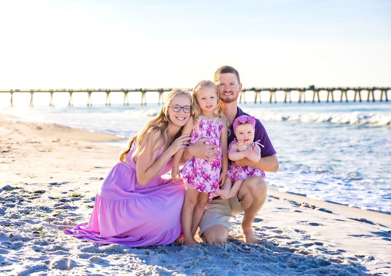 Group in coordinating pink outfits pose together on sunny beach with wooden pier in background.