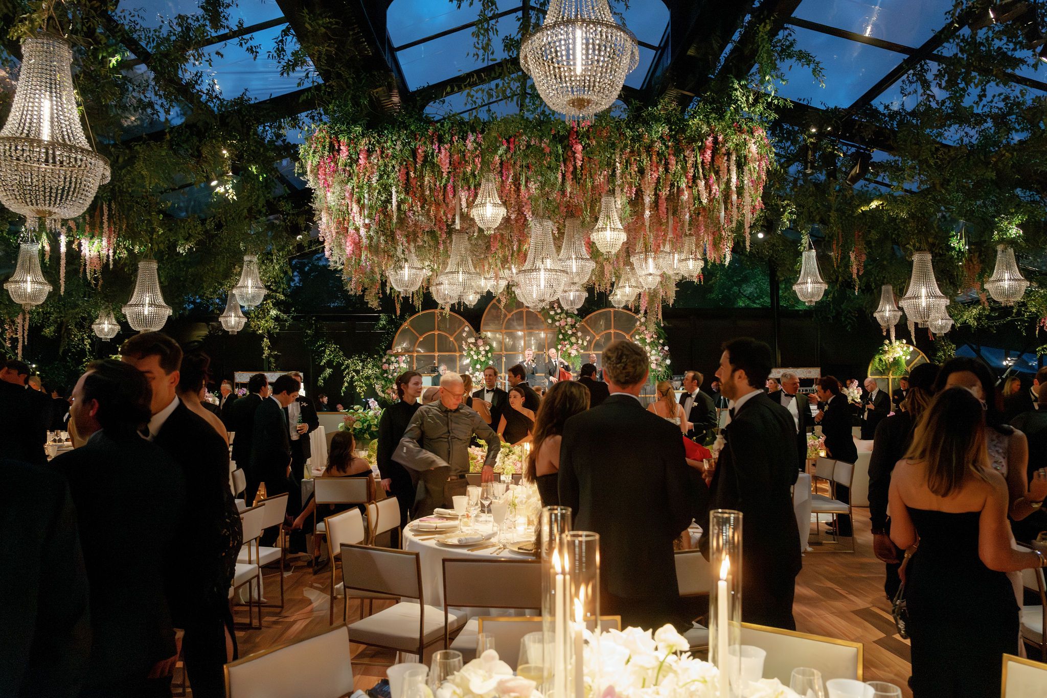 An illuminated glass greenhouse reception venue glows warmly at dusk beside a swimming pool with landscaped grounds.