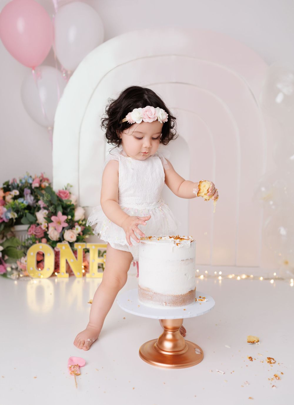 First birthday cake smash photoshoot with baby in white dress and pink balloons.