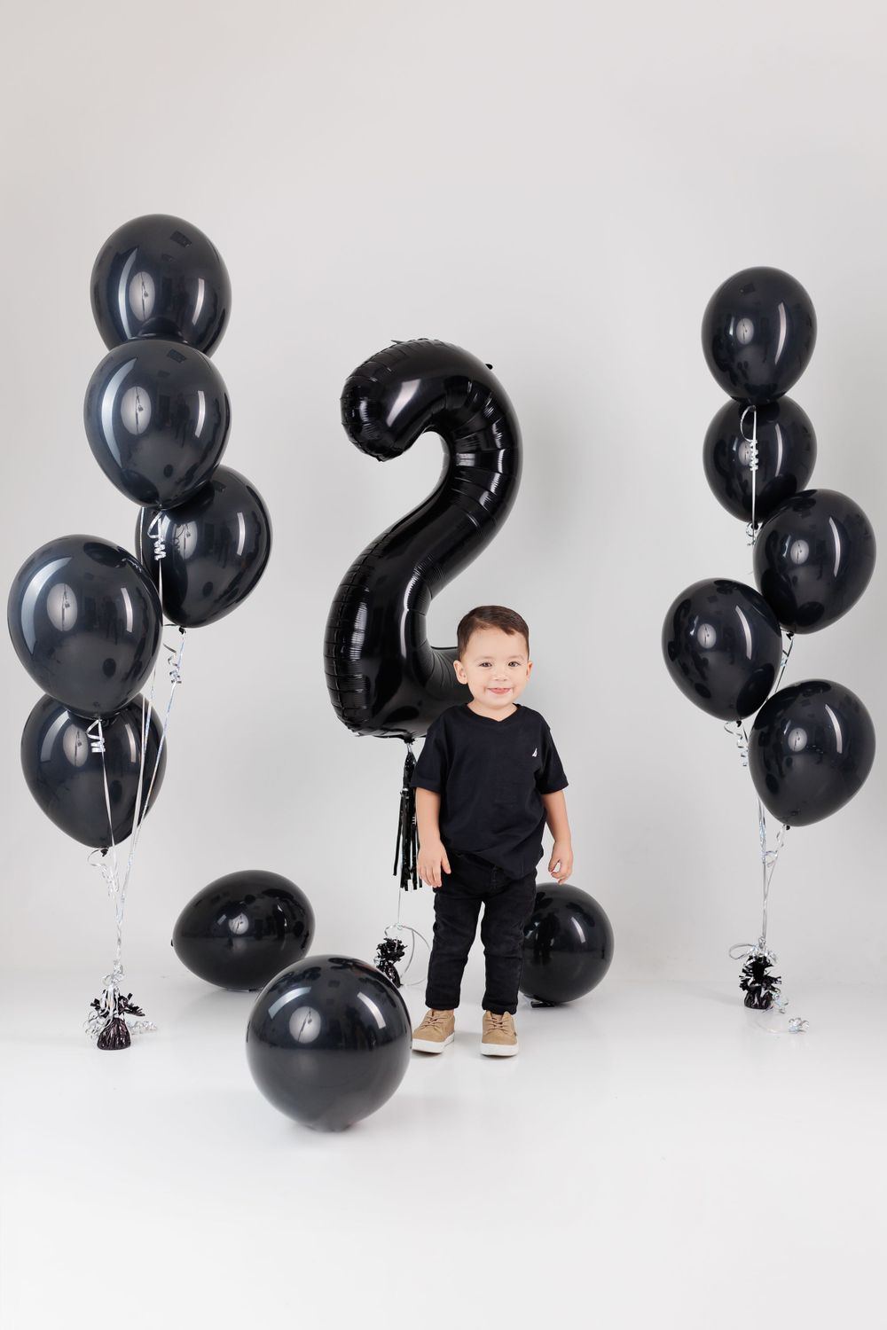 Child in black outfit plays among floating black balloons in white studio.