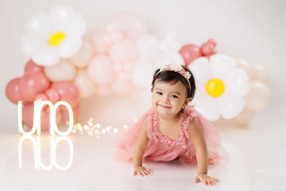 Baby in pink dress crawling by floral balloon display with illuminated 'uno' sign.