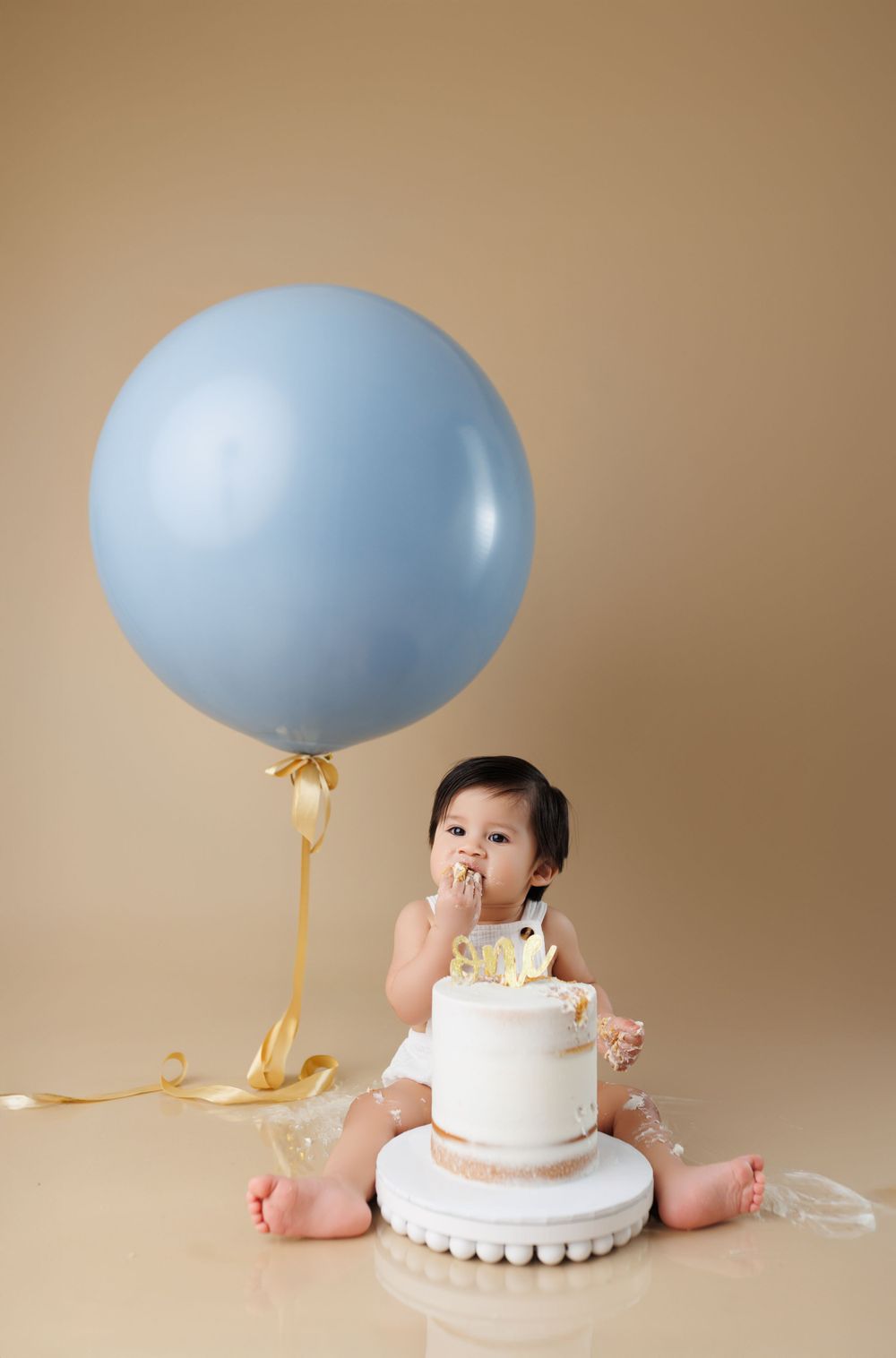 Baby enjoying birthday cake next to large blue balloon on beige background.