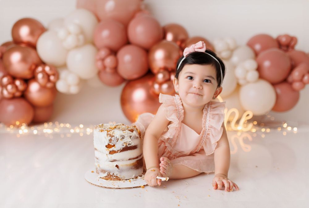 Baby next to birthday cake with rose gold balloon arch and fairy lights in background.