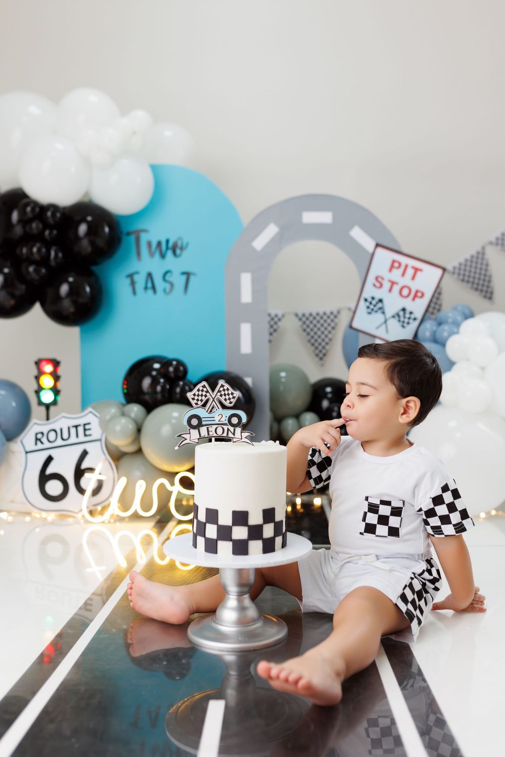 A racing themed birthday setup with a checkered cake, Route 66 sign, and balloon decorations on a gray backdrop.