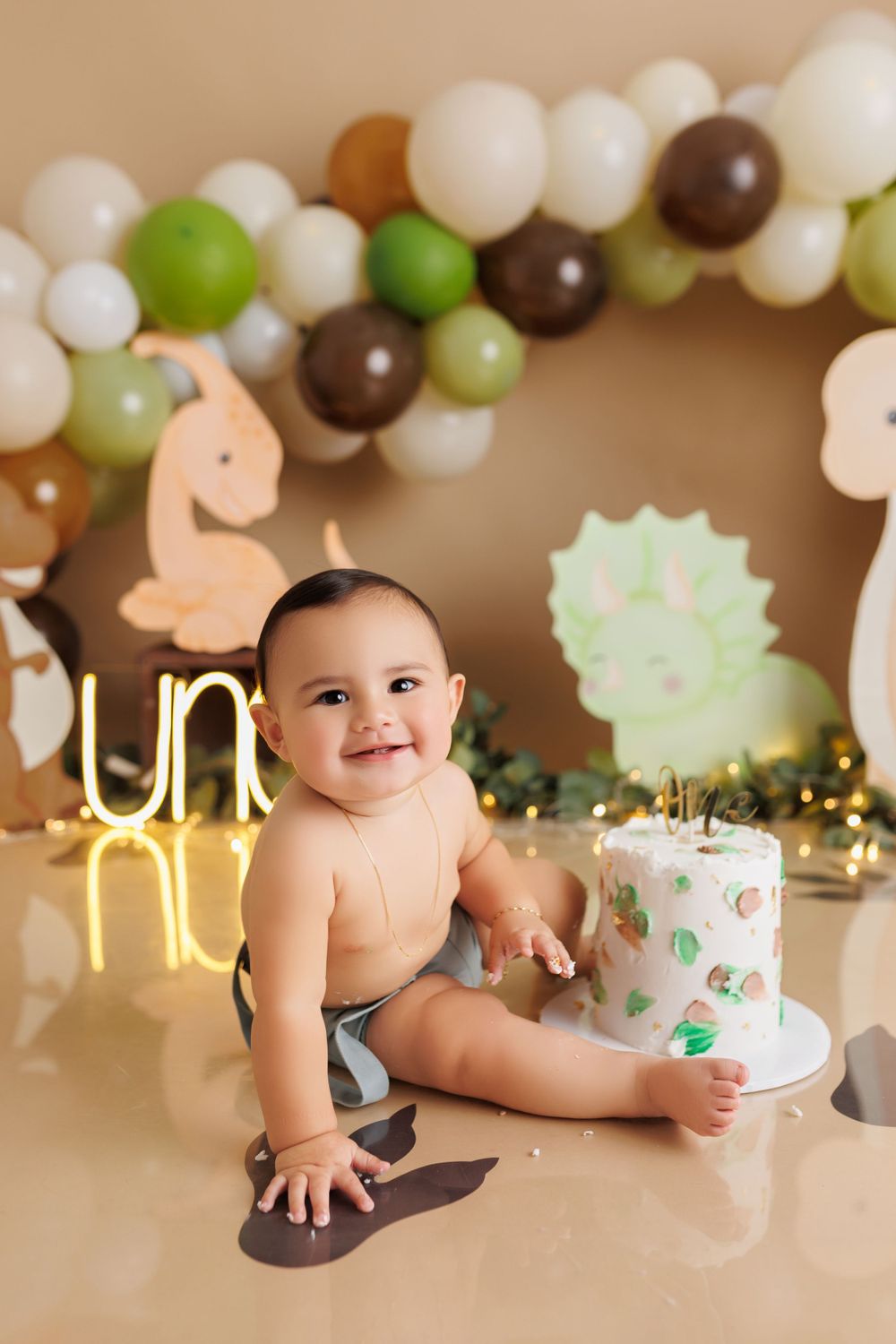 Baby sits near white cake with safari-themed balloon decorations and lighting.