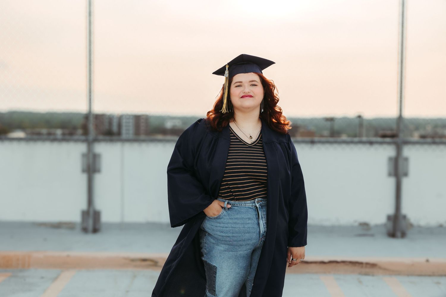 Graduate in a cap and gown stands on rooftop against a city backdrop during sunset in downtown Ann Arbor, Michigan