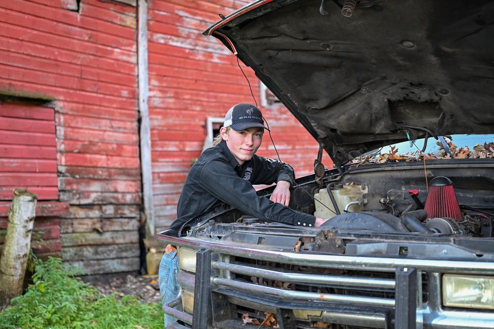 A young man leans over the engine of an old truck parked beside a weathered red barn.