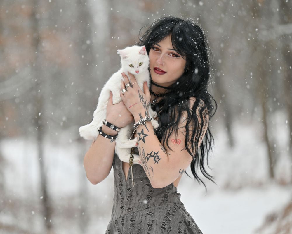 Goth woman with tattoos holding white cat in snowy winter forest wearing gray dress.