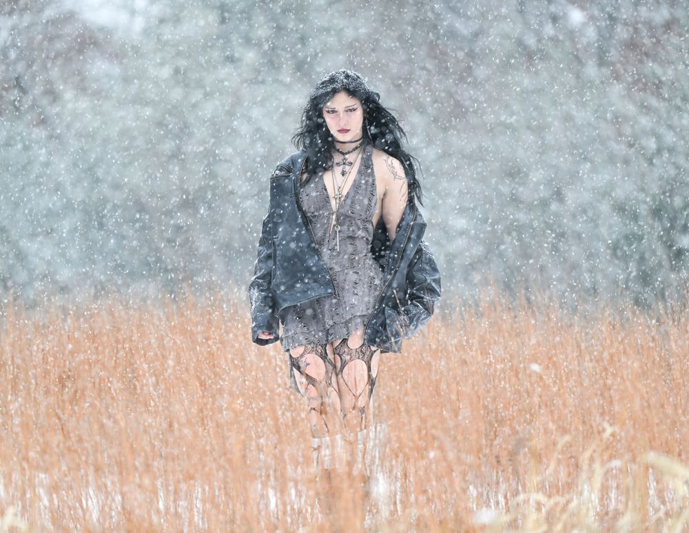 Gothic woman in gray dress standing in snow-covered field during blizzard.