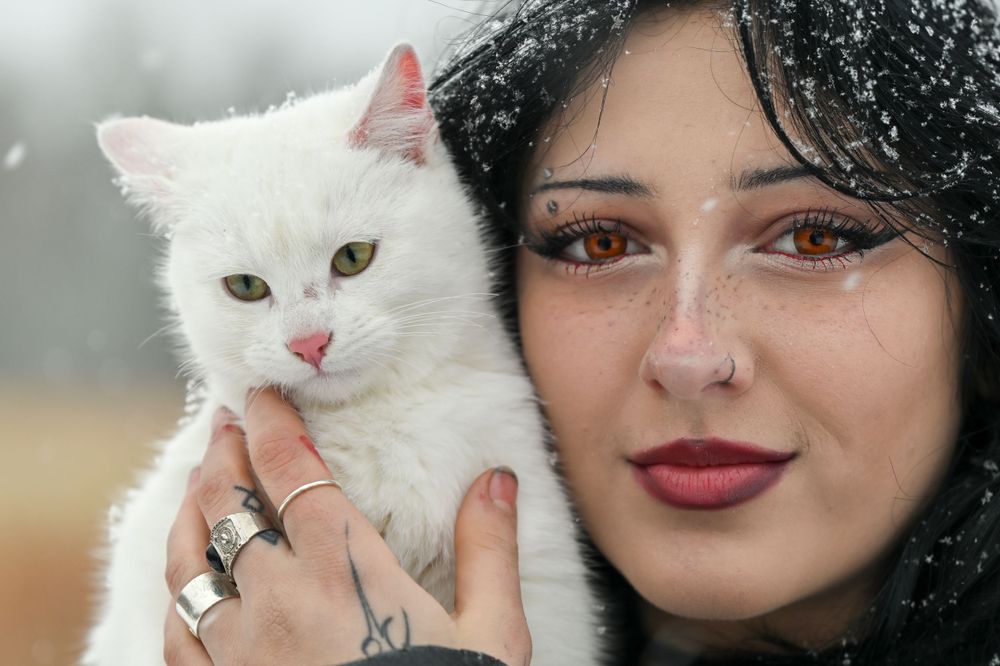 Close-up portrait of dark-haired woman with brown eyes holding white cat in winter snow.