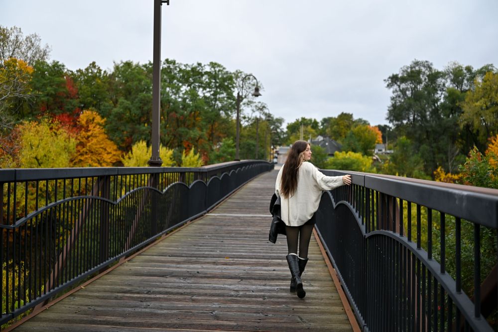 Woman in white oversized sweater strolling across wooden bridge amid stunning autumn tree colors.
