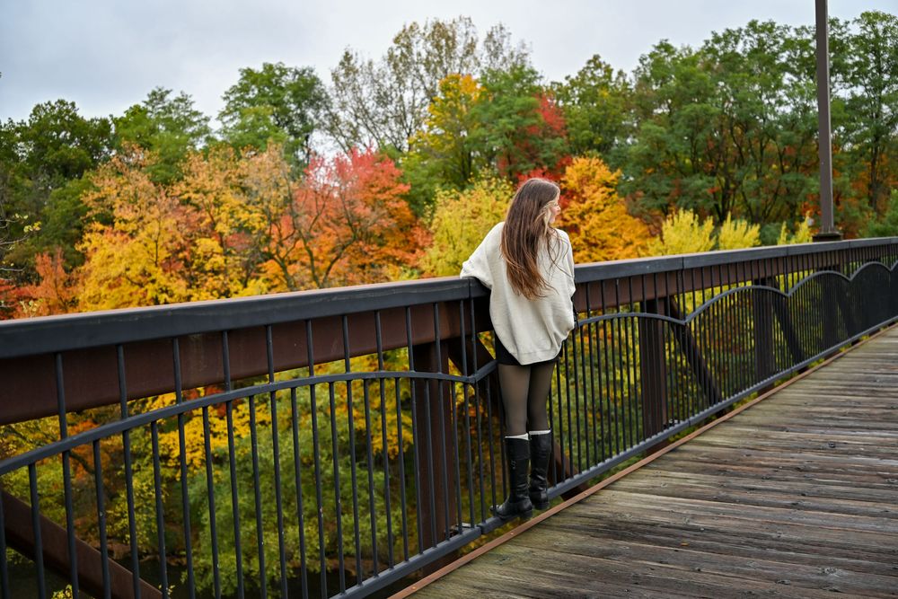 Young woman leaning on bridge railing admiring vibrant fall foliage in golden autumn colors.