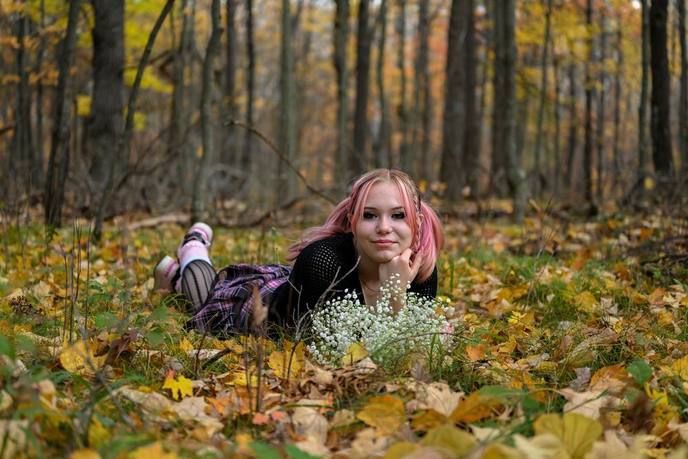 Smiling young woman with pink hair lying among colorful fall leaves in a woodland setting.
