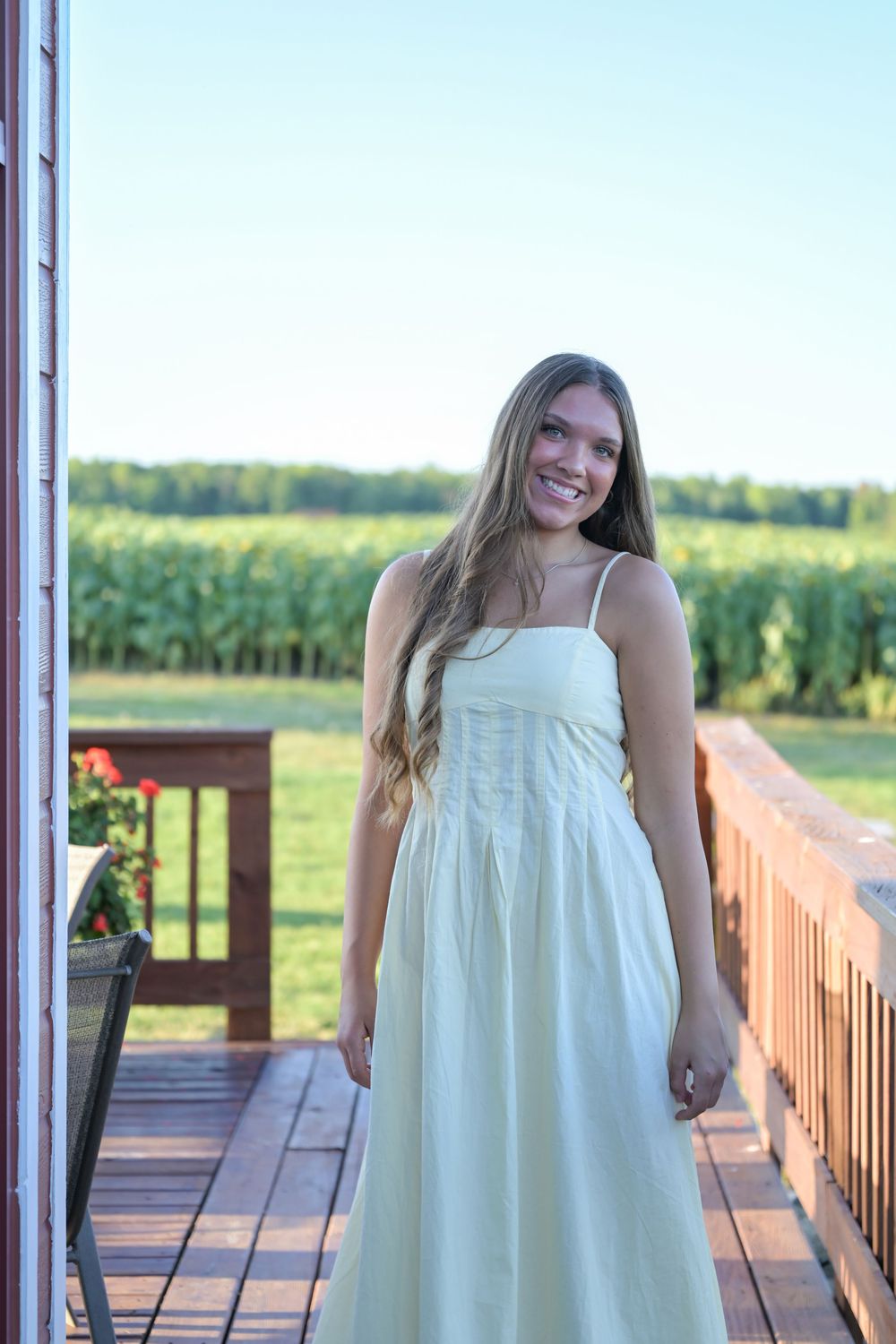 Smiling young woman in white sundress standing on wooden deck with green cornfield background on sunny day.