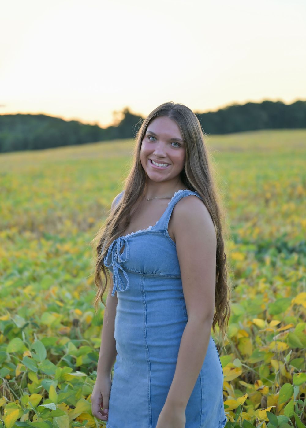 Young woman in denim dress smiling in a sunny green field at golden hour with trees in background.