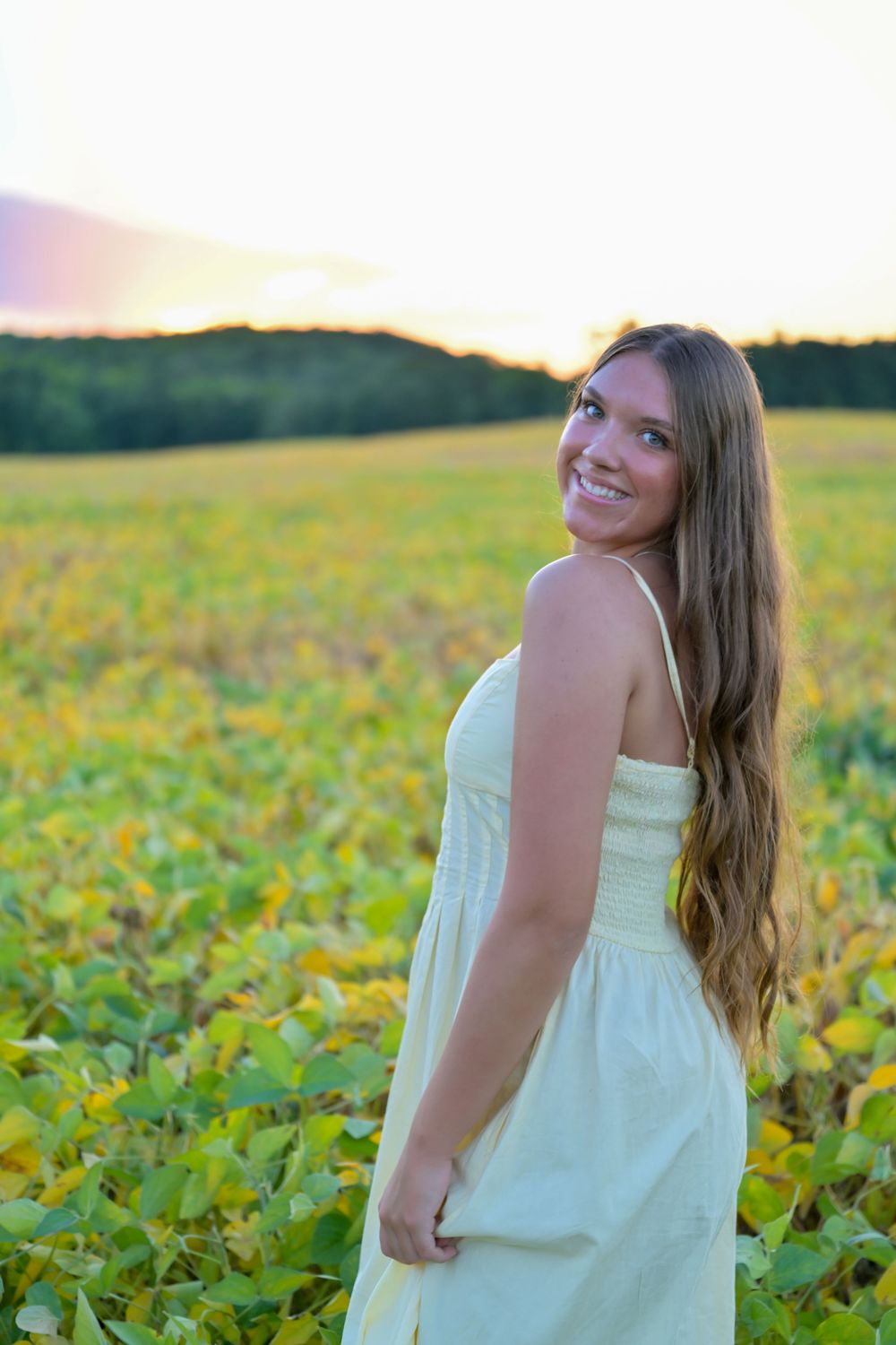 Smiling young woman in white sundress standing in a lush green field at golden hour sunset.