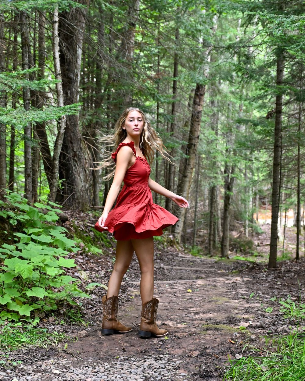 Young woman in red dress and cowboy boots twirling on a forest path surrounded by tall pine trees.