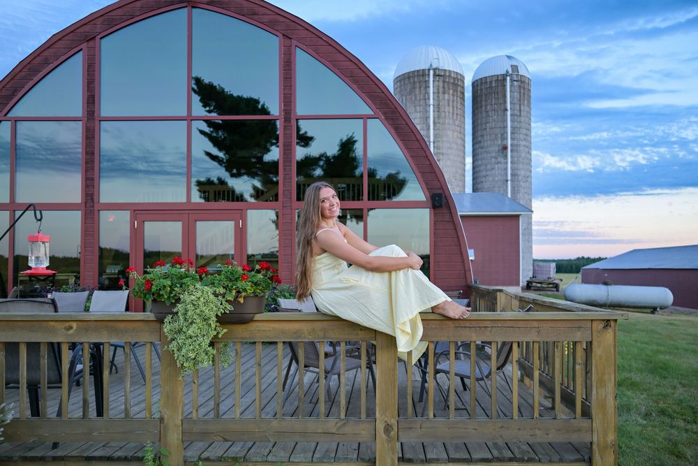 Woman in white dress sitting on wooden deck of a quonset barn with silos at sunset on a farm.
