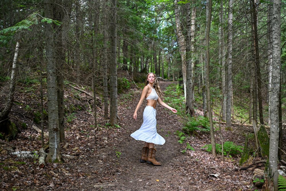 Blonde woman in white skirt and boots spinning joyfully on a scenic woodland dirt path.